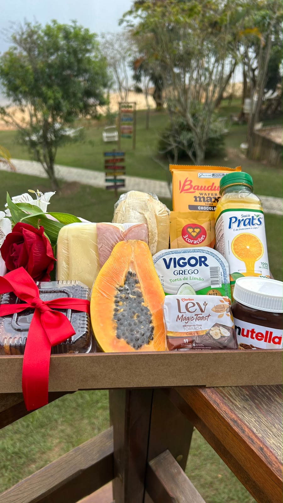 Cesta de café da manhã com frutas, frios, pães, suco, iogurte e uma rosa vermelha com laço